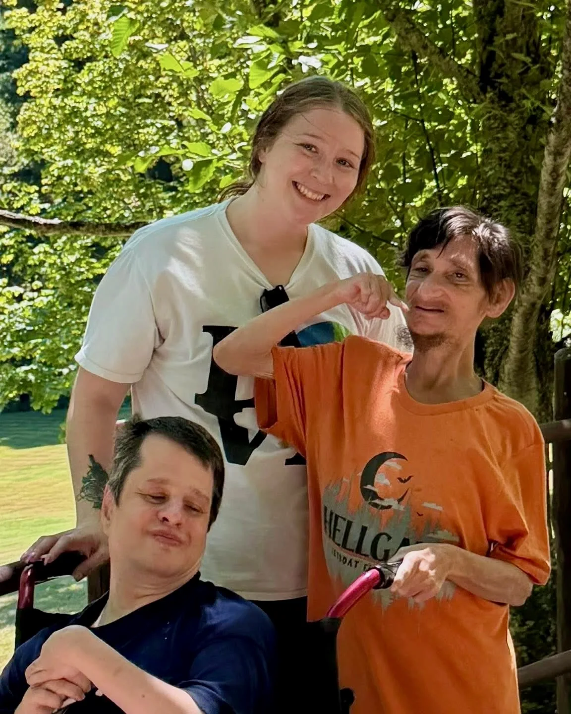A Living Opportunities DSP smiles with two men she supports outdoors on a sunny day, surrounded by green trees.