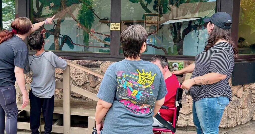DSPs and people they support look at an animal exhibit together at the zoo, with one DSP pointing out something through the window.