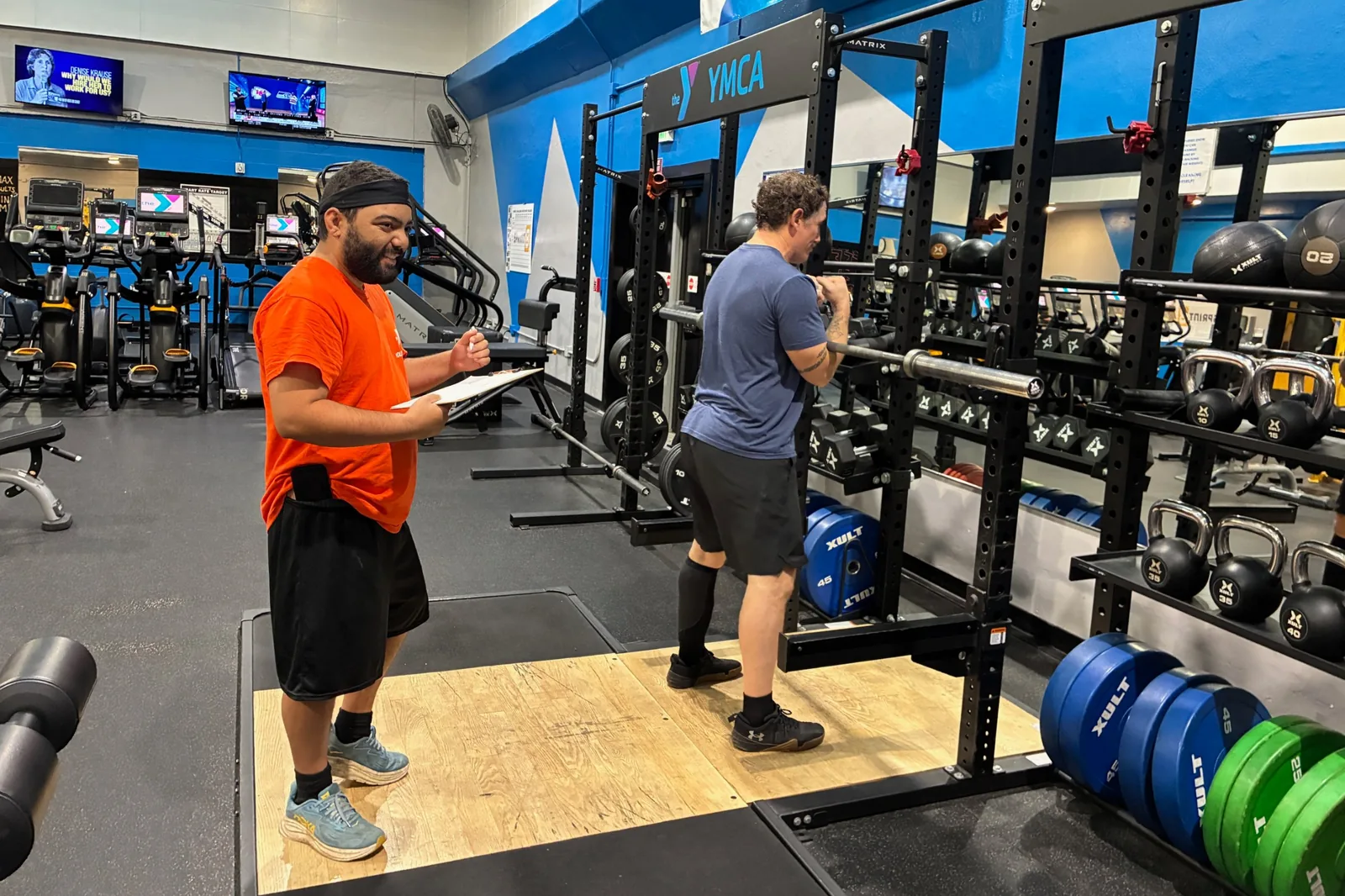 Louie Speice holds a clipboard and smiles while coaching Adam McMenomy at a squat rack in the Medford YMCA weight room.