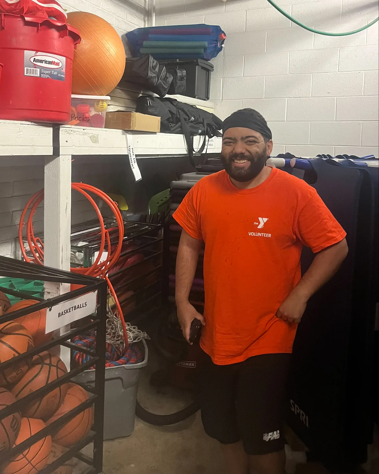 Louie Speice smiles in a YMCA volunteer shirt beside shelves of sports equipment.