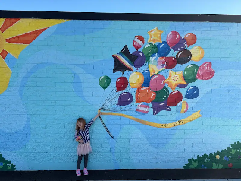 Bonnie smiling in front of a colorful community mural with painted balloons on the Living Opportunities building.
