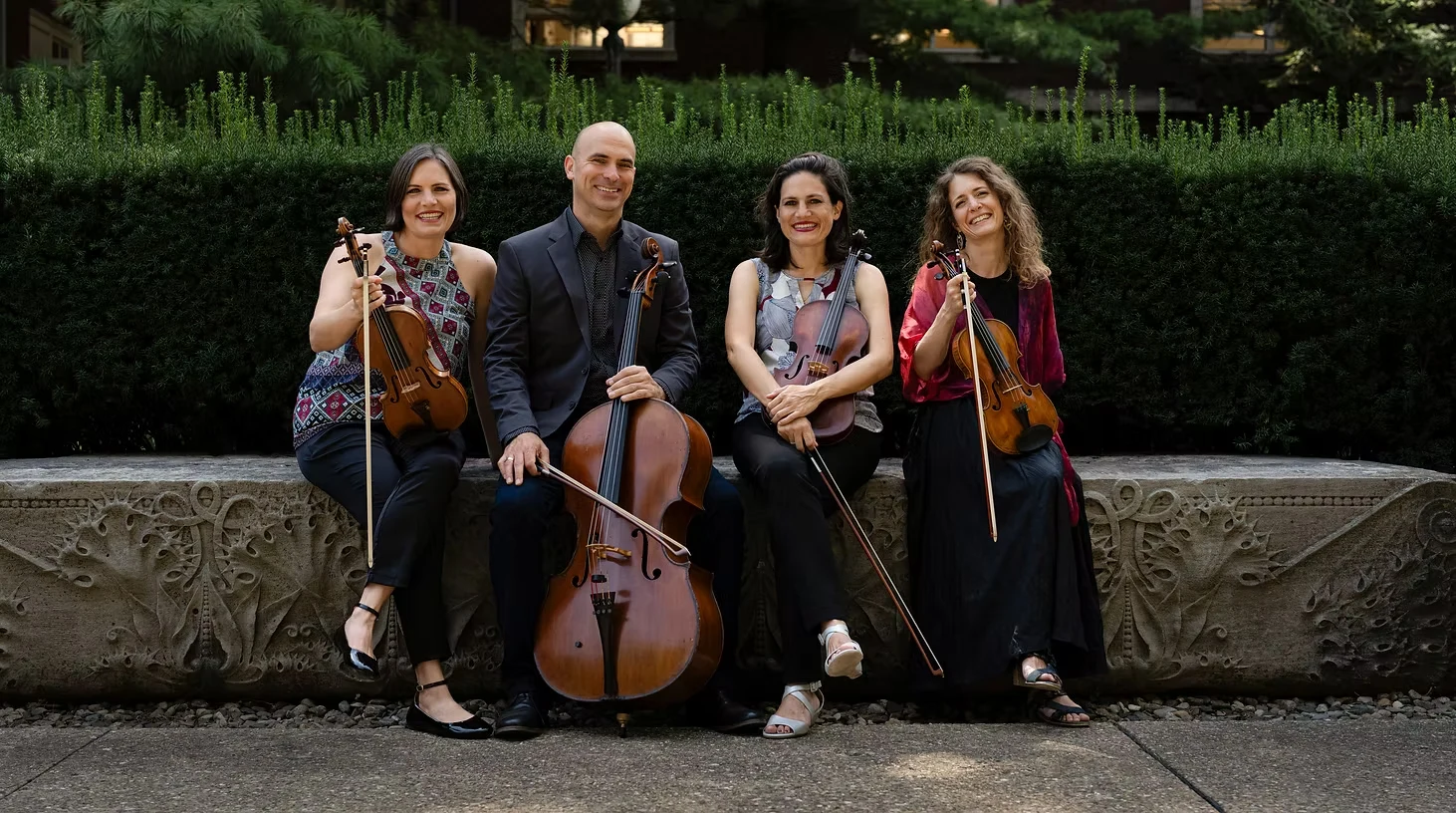 The Jupiter String Quartet seated outdoors on a carved stone bench with their violins, viola and cello