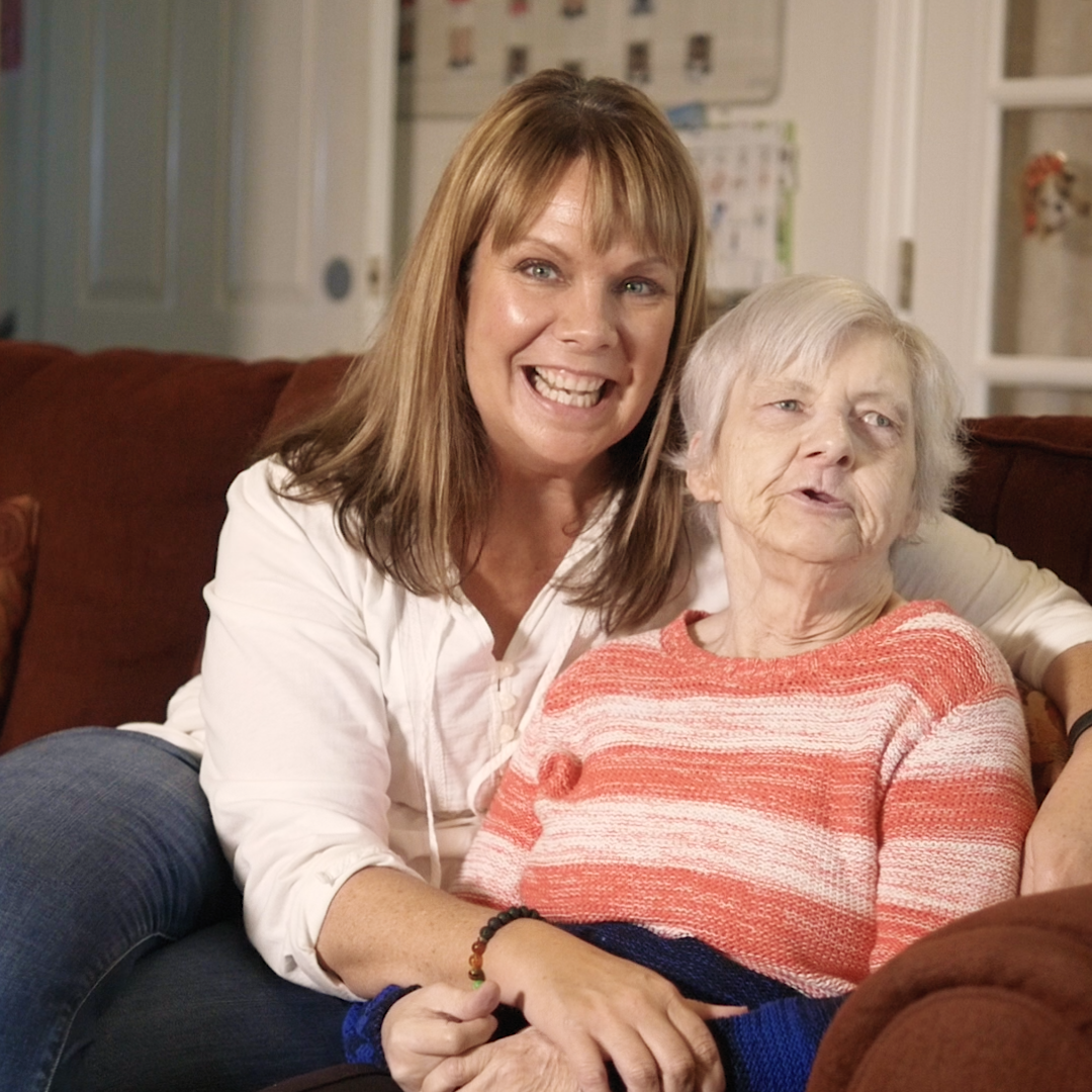 A woman sitting in a supportive position with arms around an elderly woman.
