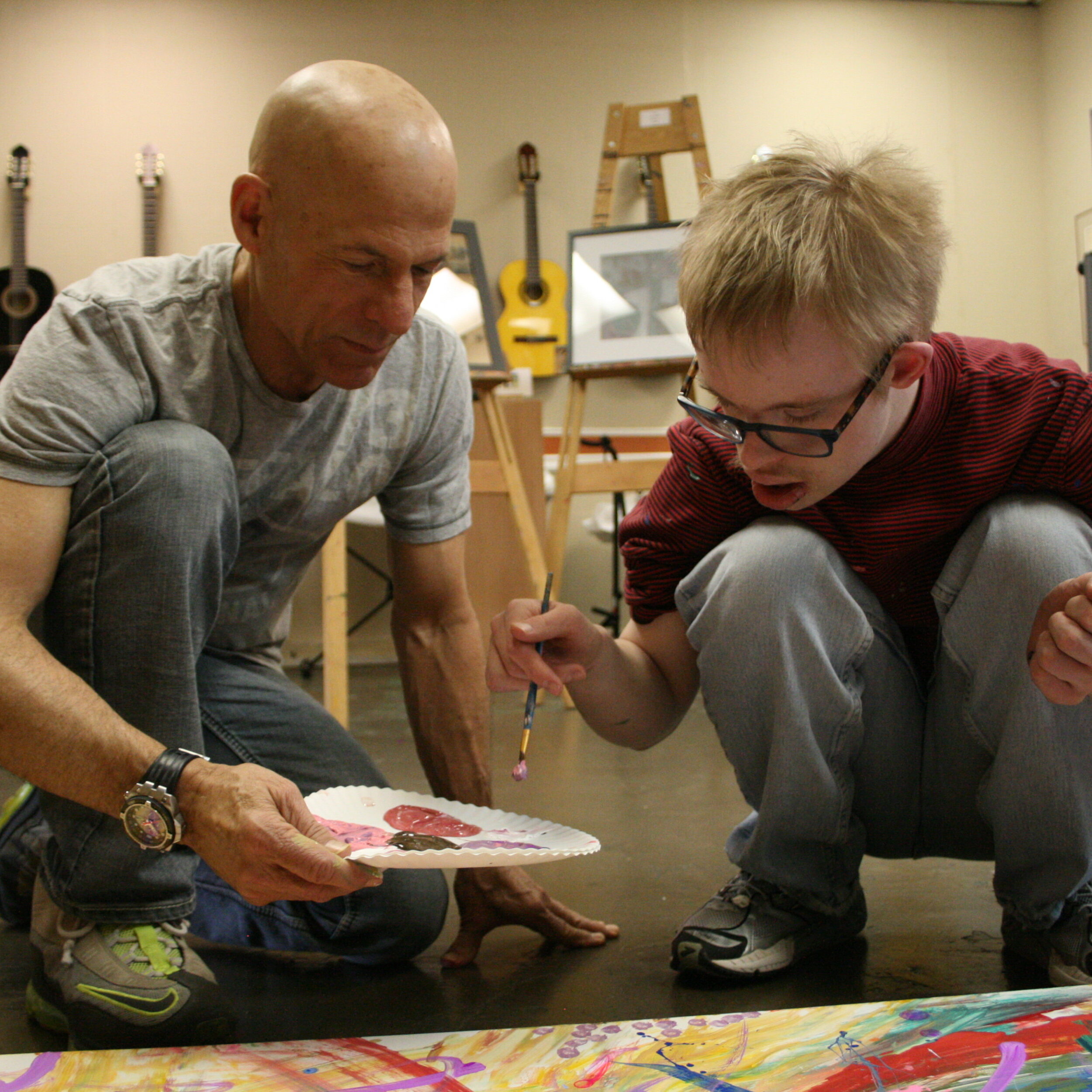A man and a young boy are crouched on the ground of an art studio working on a painting together.