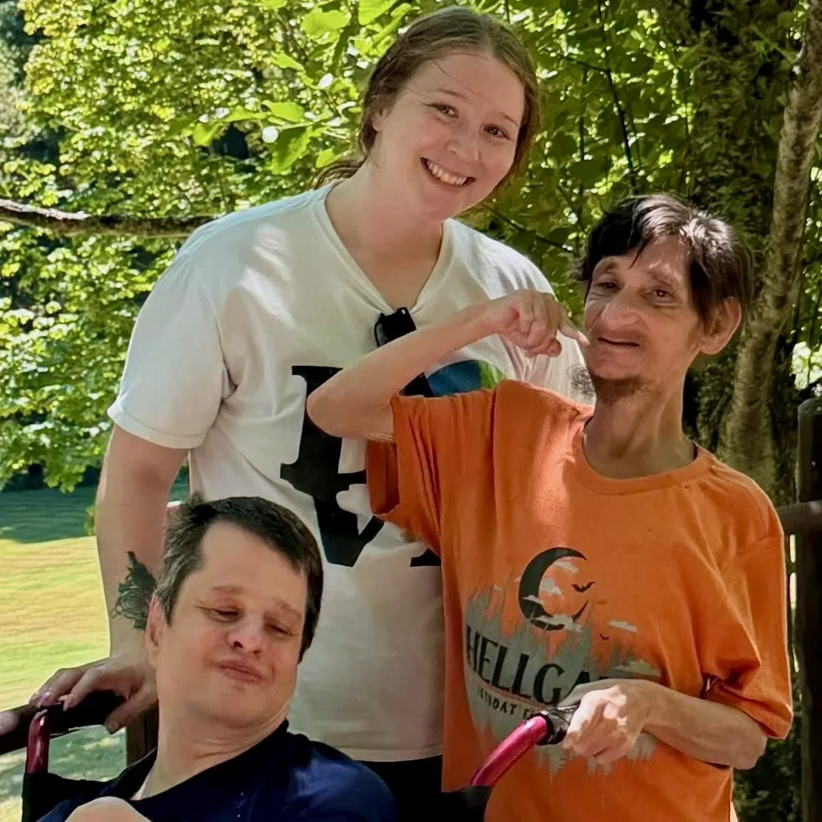 A Living Opportunities DSP smiles with two men she supports outdoors on a sunny day, surrounded by green trees.