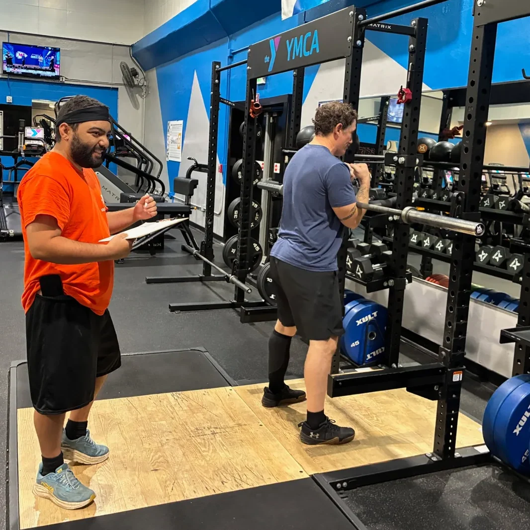 Louie Spiece holds a clipboard and smiles while coaching instructor Adam McMenomy at a squat rack in the Medford YMCA weight room.