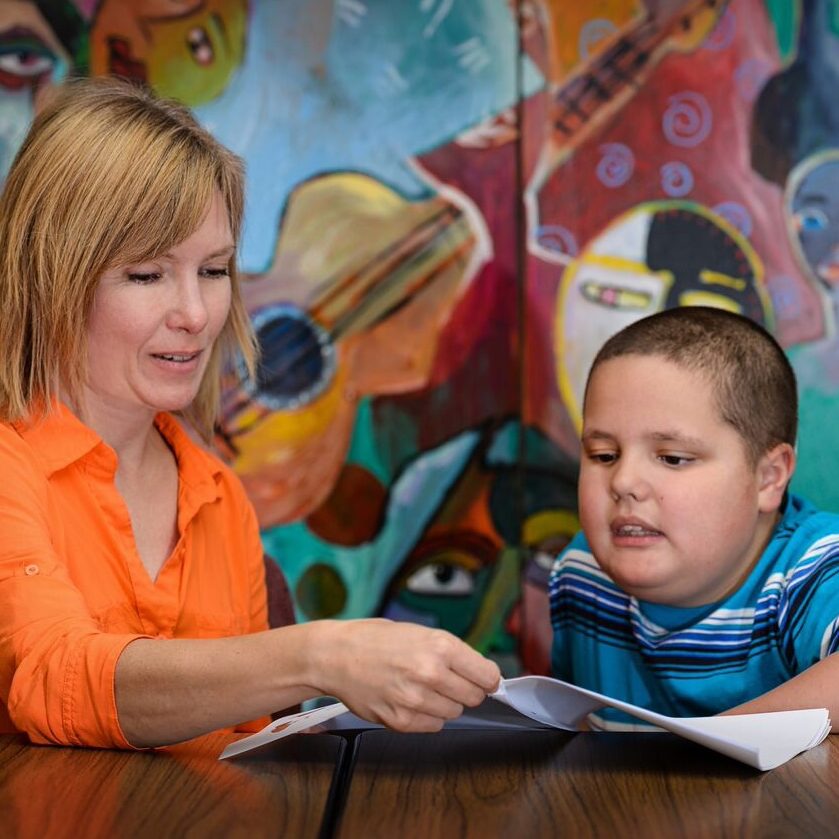 A woman is holding a book open for a youngster as they sit at a table together.